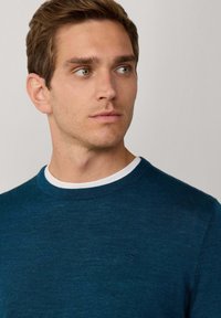 Niebieski sweter dzianinowy z okrągłym dekoltem i białymi wykończeniami wewnętrznymi, z delikatnym logo na piersi. Gładka faktura, luźny krój.