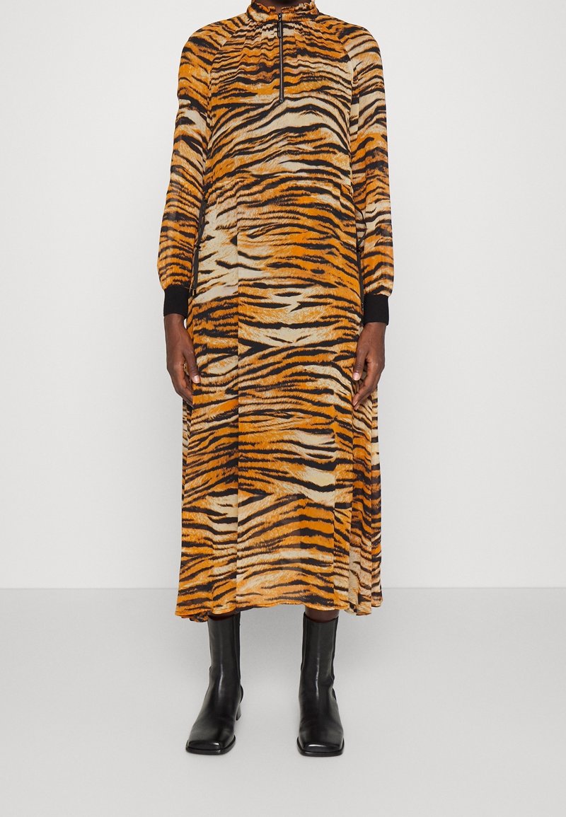 Femme portant une robe midi à manches longues imprimée tigre avec une fermeture éclair à l'avant, associée à des bottines noires, sur un fond blanc uni.