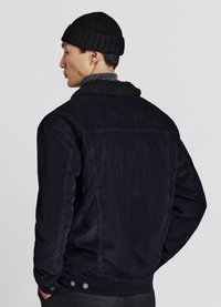 Mann, der eine schwarze gerippte Mütze und eine dunkle Cordjacke mit einem schwarzen Lammfellkragen trägt, von hinten vor einem schlichten Hintergrund gesehen.