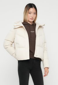 Jonge vrouw in een crèmekleurige pufferjack over een bruine hoodie en zwarte broek, staand tegen een effen witte achtergrond.