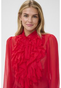 Femme blonde aux yeux bleus portant des boucles d'oreilles créoles en or et un chemisier rouge vif avec des volants à l'avant, souriant légèrement.