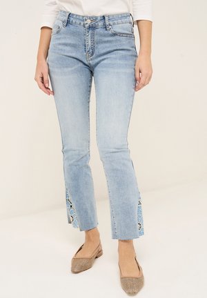 Jean bleu clair avec des fentes latérales à motif floral, porté avec des chaussures plates beiges texturées et un haut blanc à manches longues.