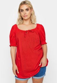 Rote Bluse mit kurzen Ärmeln, quadratischem Ausschnitt, Bindedetail in der Mitte, strukturierter Stoff und puffigen Ärmeln. Kombiniert mit Jeansshorts.