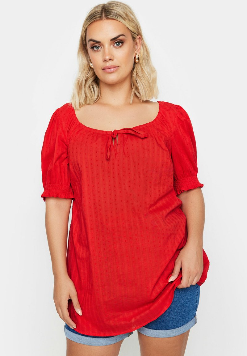 Rote Bluse mit kurzen Ärmeln, quadratischem Ausschnitt, Bindedetail in der Mitte, strukturierter Stoff und puffigen Ärmeln. Kombiniert mit Jeansshorts.