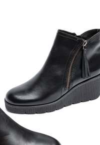 Black leather ankle boots with a wedge sole, featuring a side zip and tassel detail. Smooth finish and textured, ridged sole.