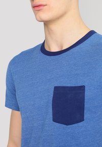 Niebieska koszulka z krótkim rękawem i okrągłym granatowym dekoltem oraz granatową kieszonką po lewej stronie klatki piersiowej. Materiał wydaje się miękki i ma melanżową teksturę.