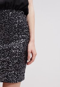 Jupe noire à sequins avec une coupe ajustée. Présente une texture scintillante et une forme mettant en valeur la taille. Main visible portant un chemisier.