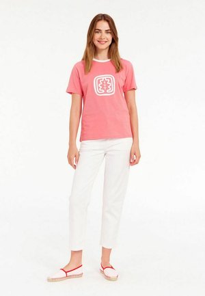 T-shirt en coton rose à manches courtes et col rond blanc, avec un logo graphique blanc sur le devant. Associé à un pantalon blanc et des espadrilles rouges.