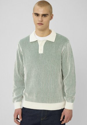 Jeune homme aux cheveux blonds courts portant un pull en maille à rayures verticales blanches et vertes par-dessus une chemise blanche et un jean bleu foncé, debout devant un fond uni.