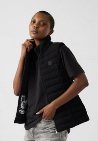 Gilet imbottito nero con colletto alto, chiusura con zip e finitura testurizzata. Indossato sopra una t-shirt nera, con un interno a fantasia.