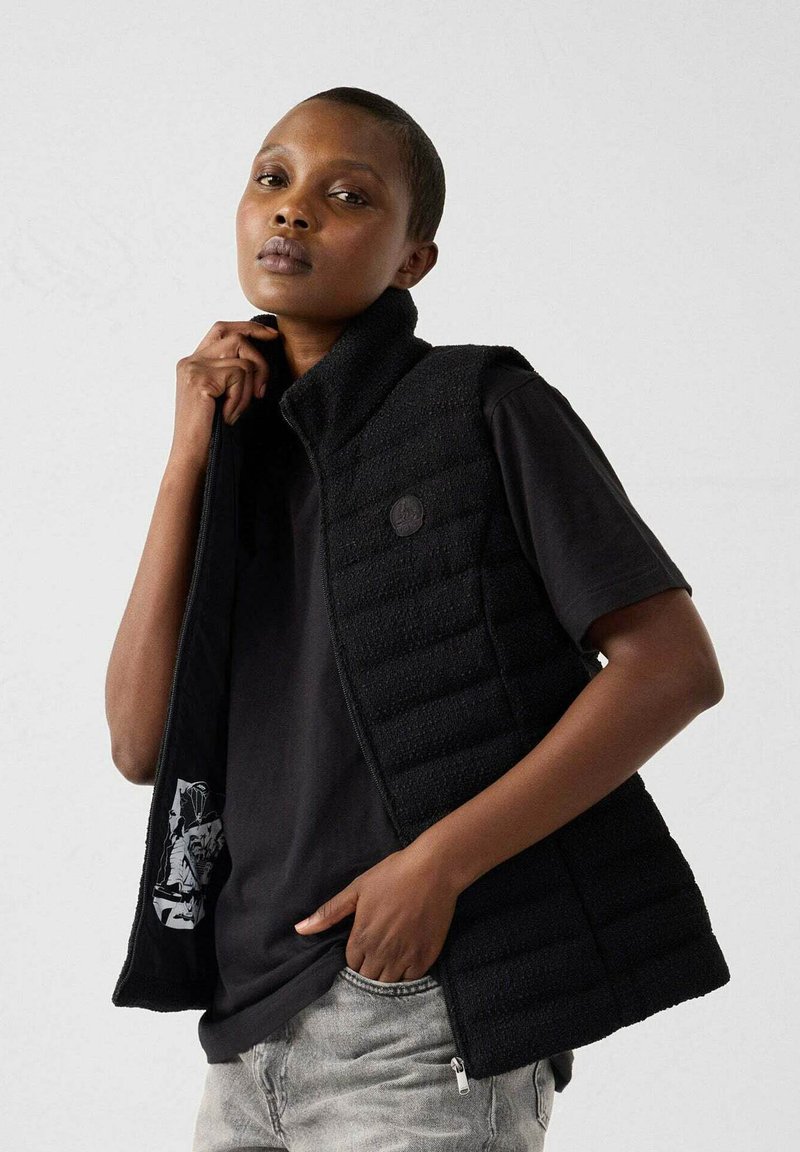 Gilet imbottito nero con colletto alto, chiusura con zip e finitura testurizzata. Indossato sopra una t-shirt nera, con un interno a fantasia.