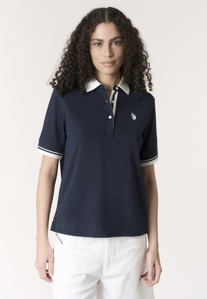 Donna con capelli ricci che indossa una polo blu navy con colletto e polsini bianchi, abbinata a pantaloni bianchi, in piedi davanti a uno sfondo neutro.