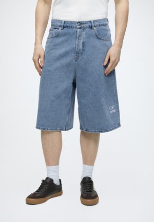 Mężczyzna w jasnoniebieskich spodenkach jeansowych do kolan, białych skarpetkach, czarno-brązowych trampkach i białej koszuli na jednolitym tle.