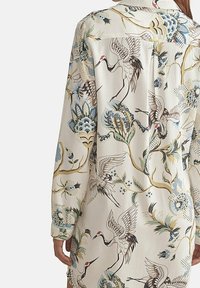 Chemise à manches longues avec un fond crème et des motifs élaborés de grues et de fleurs dans des tons de bleu, vert et or, fabriquée en tissu lisse.