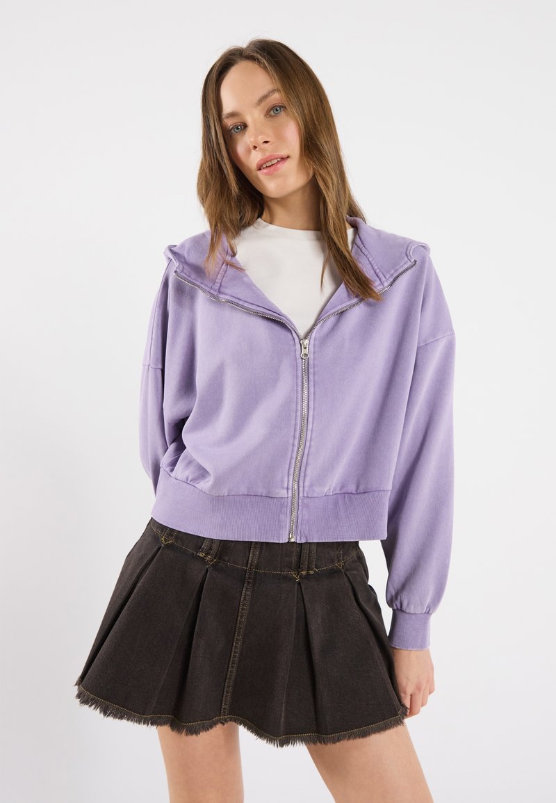 Felpa viola con zip e tasca frontale, abbinata a una gonna plissettata in denim scuro con orli sfilacciati.