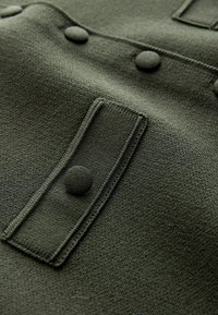 Gros plan sur un tissu vert foncé avec des boutons texturés et un détail de rabat rectangulaire cousu.