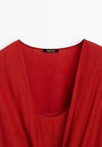 Rode kleding met een diepe v-hals en een zak, met een label waarop "Massimo Dutti" staat. Zachte stofstructuur zichtbaar.