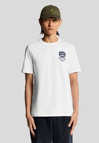 Hvid bomulds t-shirt med korte ærmer, med et navy grafisk logo på brystet. Båret med en mørkegrøn cap. Enkelt og afslappet design.