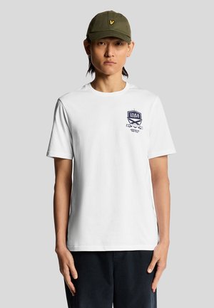 Witte katoenen t-shirt met korte mouwen, met een marineblauw grafisch logo op de borst. Ggedragen met een donkergroene pet. Eenvoudig en casual ontwerp.