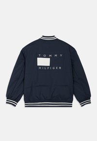 Chaqueta bomber azul marino con cuello y puños de rayas blancas acanaladas. Presenta un parche rectangular con el texto "TOMMY HILFIGER".