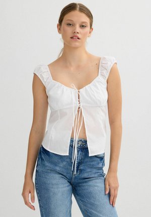 Joven mujer con una blusa blanca sin mangas con detalles de nudo en el frente y jeans de tiro alto azules, posando frente a un fondo liso.