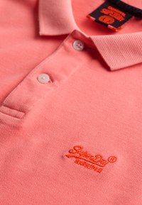 Polo rose corail avec deux boutons blancs, col plié et logo de marque brodé orange sur la poitrine.