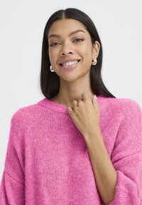 Jeune femme aux longs cheveux noirs portant des boucles d'oreilles en argent et un pull en tricot rose vif, souriant doucement avec une main près de son collarbone.