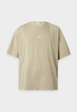 Jeune homme noir aux cheveux tressés portant un t-shirt beige oversize et un pantalon cargo vert olive, debout devant un fond blanc uni.