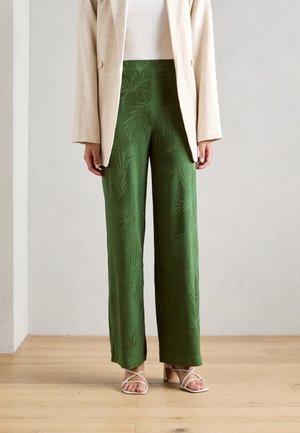 Femme portant un pantalon vert taille haute à jambes larges avec un motif subtil de feuilles, un blazer beige, un haut blanc et des sandales beiges à brides, debout sur un sol en bois.