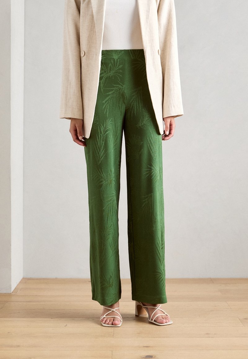 Femme portant un pantalon vert taille haute à jambes larges avec un motif subtil de feuilles, un blazer beige, un haut blanc et des sandales beiges à brides, debout sur un sol en bois.