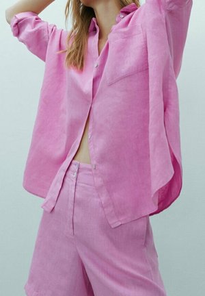 Femme portant une chemise ample rose à boutons et un short taille haute assorti, les bras levés, sur un fond clair uni.