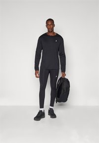 Svart långärmad skjorta och åtsittande leggings, båda gjorda av slät tyg. Svarta sportskor och en svart ryggsäck ingår.