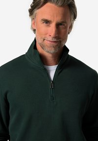 Musta vihreä vetoketjupulleni. Tikkipinta, jossa on korkea kaulus ja hopeanvärinen vetoketju. Vasemmassa rinnassa on hillitty logo.