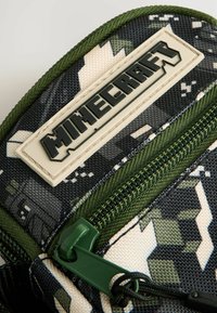 Camouflage-mønsteret lynlåspose i nuancer af grøn og sort, der pryder et hævet hvidt "Minecraft" logo og struktureret stof.