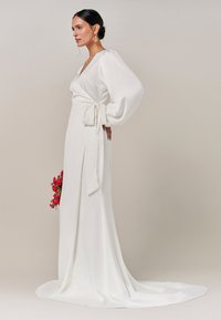 Weißes Wickelkleid mit langen, puffigen Ärmeln, tiefem V-Ausschnitt, mit einem Band an der Taille. Fließender Stoff mit einem leichten Zug; Model hält pinkfarbene Blumen.