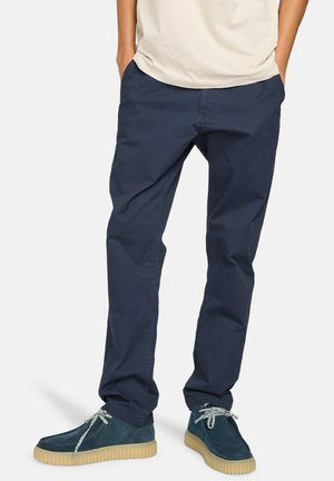 Navy blue cotton chinos, straight fit with side pockets, paired with green suede shoes featuring gray laces and a textured rubber sole.