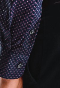 Rękaw granatowej koszuli z różowo-białym wzorem w kropki i dwoma widocznymi guzikami, obok czarnego materiału i nagiego przedramienia.