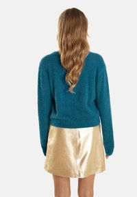 Pulover fuzos de culoare teal, cu mâneci lungi, asortat cu o fustă mini metalică de aur lucios. Textură moale și croială lejeră, fără modele vizibile.