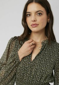 Blouse florale verte avec des manches longues transparentes, un décolleté en V et une taille smockée. Un petit motif de fleurs blanches et violettes ajoute du détail.