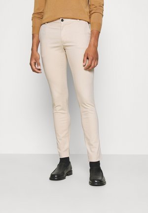Person trägt beigefarbene Slim-Fit-Hose, schwarze Socken, schwarze Lederschuhe und ein braunes Langarm-Oberteil und steht vor einem einfarbigen Hintergrund.
