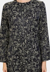 Robe noire à manches longues et encolure ronde, présentant un motif floral dans des tons crème et or sur un tissu texturé.