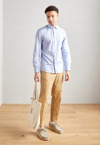 Lichtblauw overhemd met knopen, beige linnen broek, grijze suède schoenen en een canvas tote bag. Houten vloer, neutrale wandachtergrond.