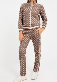Brown zip-up jacket and matching pants with white floral pattern, striped accents, and elastic cuffs. Worn with white sneakers.