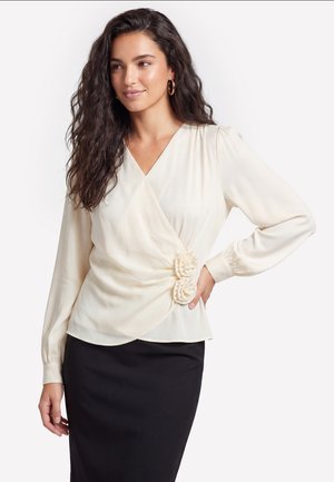 Frau mit langen lockigen dunklen Haaren, die eine cremefarbene Wickelbluse mit Blumendetails und einen schwarzen Rock trägt, steht mit der Hand auf der Hüfte.