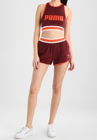 Camiseta deportiva corta color burdeos con detalles en blanco y naranja, combinada con pantalones cortos a juego en burdeos con un ribete naranja. Zapatillas blancas.