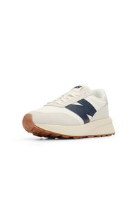 Cream-colored sneaker with navy suede accents, textured upper, and a rubber gum sole. Features a round toe and lace-up design.