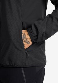 Svart jacka med texturerat mönster, med en dragkedjeförsedd sidoficka och elastiska ärmslut. Tillverkad av ett lättviktigt, flexibelt material.