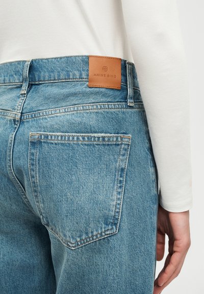 Close-up of blue denim jeans back pocket and brown leather Anine Bing brand label, worn with white long sleeve top.