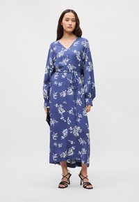 Μακρύ μπλε φόρεμα με λευκό λουλουδάτο μοτίβο, v-ντεκολτέ, μακριά μανίκια, ζώνη στη μέση και πλαϊνές σχισμές, συνδυασμένο με μαύρες στιλέτο σάντες.