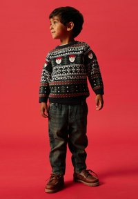 Bambino che indossa un maglione di Natale nero con motivi di Babbo Natale e fiocchi di neve, abbinato a jeans blu in denim e scarpe marroni con lacci.